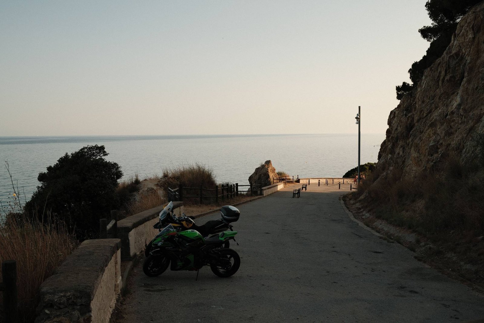 Moto garée près d’un muret de pierre sur une route tranquille face à la mer, entourée de falaises — le type de paysage serein qui caractérise une bonne partie de la côte de Menorca