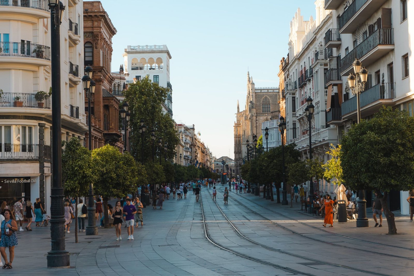Amplia avenida peatonal del centro de Sevilla con la torre de la Giralda al fondo y las vías del tranvía, en un atardecer de verano